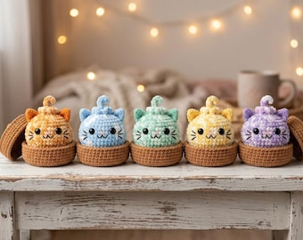 May include: Five small, round, crocheted cat figures in orange, blue, green, yellow, and purple, each nestled inside a brown crocheted base. The cats feature pointed ears, black embroidered eyes, whiskers, and a small swirl on top of their heads.