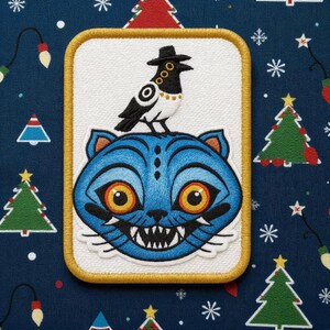 May include: Embroidered patch featuring a blue cat with orange eyes and a black bird wearing a hat. The patch has a white background with a gold border. The background is a dark blue fabric with Christmas tree and star patterns.