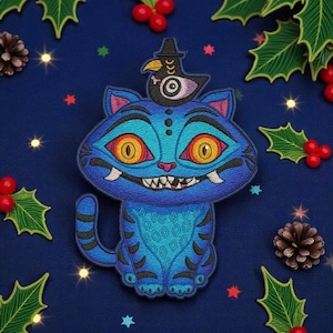 May include: A blue cat patch with a bird wearing a hat on its head. The cat has large yellow eyes, sharp teeth, and a patterned design. The patch is surrounded by holly and berries on a dark blue background.