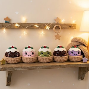 May include: Five small, round, crocheted plush toys shaped like Christmas puddings, each sitting in a tan crocheted bowl. The puddings are dark brown, light pink, light green, medium brown, and light purple, all with white frosting tops.