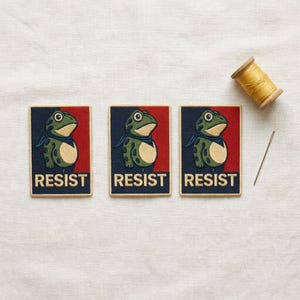 May include: Three rectangular embroidered patches featuring a green frog with a blue neckerchief against a split dark blue and red background. The word RESIST is printed in bold cream letters at the bottom of each patch, accompanied by a spool of thread.