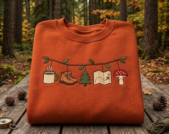 Op de afbeelding: Een roestoranje sweater met ronde hals en een geborduurd ontwerp van een slinger met bladeren waaraan een koffiemok, bruine wandelschoenen, een groene dennenboom, een open kaart met een route en een rood-witte paddenstoel hangen.