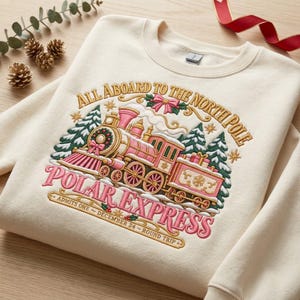 May include: A cream-colored crewneck sweatshirt featuring an embroidered design of a pink and gold steam train. The text reads ALL ABOARD TO THE NORTH POLE, POLAR EXPRESS, ADMITS ONE, DECEMBER 24, ROUND TRIP. The graphic includes green pine trees.
