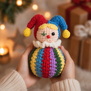 May include: A crochet jester doll featuring a round body with vertical stripes in red, blue, yellow, green, and purple. The head has a beige face, a red nose, one black eye, and one green star-shaped eye, topped with a red and blue jester hat.