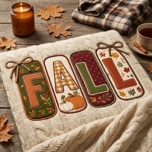 May include: A cream-colored cable knit blanket featuring four large fabric applique letters spelling FALL. Each letter is bordered by decorative stitching and adorned with autumn-themed patterns including pumpkins, leaves, and bows.