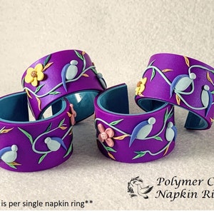 May include: Four purple polymer clay napkin rings with a floral and bird design. The rings are decorated with yellow, pink, blue, and green accents. **price is per single napkin ring**
