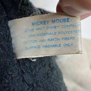 Peut inclure: Gros plan sur une &eacute;tiquette d'un jouet en peluche Mickey Mouse. L'&eacute;tiquette porte le texte bleu "MICKEY MOUSE", "THE WALT DISNEY COMPANY", "ALL NEW MATERIALS POLYESTER", "COTTON AND RAYON FIBERS" et "SURFACE WASHABLE ONLY".