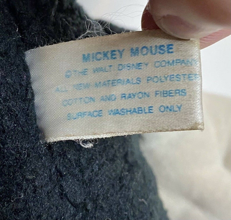 Peut inclure: Gros plan sur une &eacute;tiquette d'un jouet en peluche Mickey Mouse. L'&eacute;tiquette porte le texte bleu "MICKEY MOUSE", "THE WALT DISNEY COMPANY", "ALL NEW MATERIALS POLYESTER", "COTTON AND RAYON FIBERS" et "SURFACE WASHABLE ONLY".