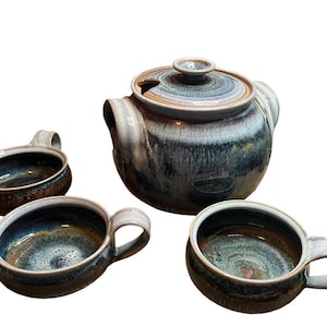 May include: A ceramic tea set with a teapot and three matching cups. The set features a speckled glaze in shades of blue, brown, and gray. The teapot has a lid and spout, while the cups have handles. A rustic, handmade aesthetic.