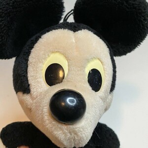 Peut inclure: Peluche Mickey Mouse avec des oreilles noires, un nez noir et des yeux jaunes. Le jouet a un visage blanc et de la fourrure noire autour des yeux et des oreilles. Personnage Disney classique.