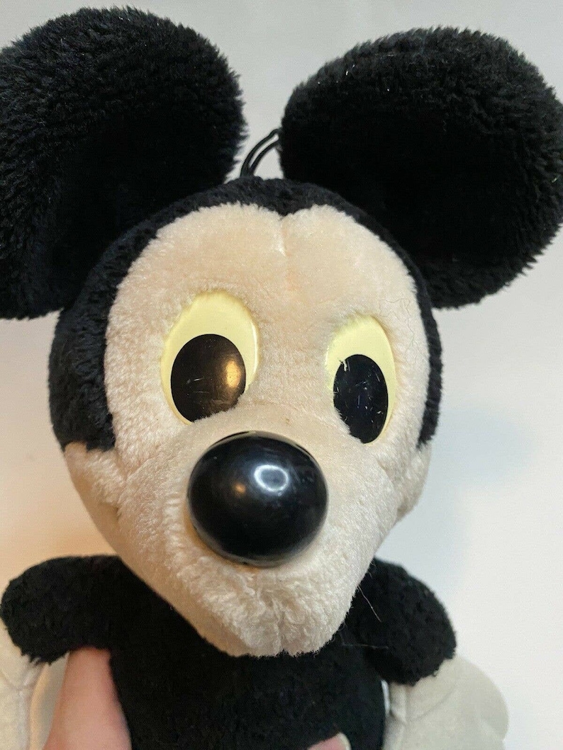 Peut inclure: Peluche Mickey Mouse avec des oreilles noires, un nez noir et des yeux jaunes. Le jouet a un visage blanc et de la fourrure noire autour des yeux et des oreilles. Personnage Disney classique.