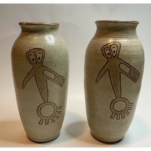 Puede incluir: Dos jarrones de cerámica beige moteada con una figura marrón estilizada grabada en la parte delantera. La figura tiene cabeza, brazos y una base circular con un diseño similar al sol. Los jarrones tienen un borde ligeramente acampanado.