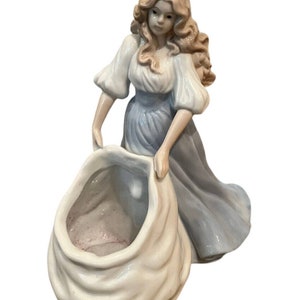 May include: A porcelain figurine of a woman in a long dress, holding a large, open sack. The woman has long, wavy blonde hair and is wearing a white blouse and a light blue dress. The figurine is detailed and elegant.