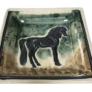Może przedstawiać: Kwadratowy ceramiczny talerz z ilustracją czarnego konia. Talerz ma zielono-brązowy krajobraz z napisem "JENNIFER & COWBOY" u góry. Ma czarną obwódkę i kremową podstawę.