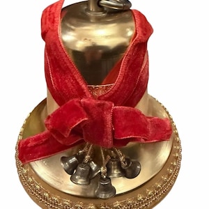 May include: A gold-colored bell with a red velvet ribbon tied in a bow. Small silver bells hang from the bow. The bell sits on a gold-colored base with a decorative edge. A ring is attached to the top of the bell.