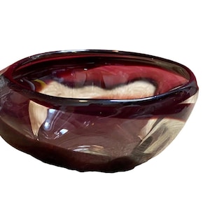 Op de afbeelding: Een handgeblazen glazen kom met een unieke, organische vorm. De kom heeft een diepe bordeauxrode kleur met wervelende witte en heldere accenten. De rand heeft een ongelijke, getextureerde rand. Deze decoratieve kom is ideaal voor presentatie of serveren.