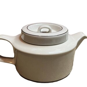 May include: A cream-colored ceramic teapot with a matching lid. The teapot has a rounded body, a curved handle, and a spout. The lid is white with a black trim and a small handle. The teapot is designed for brewing tea.