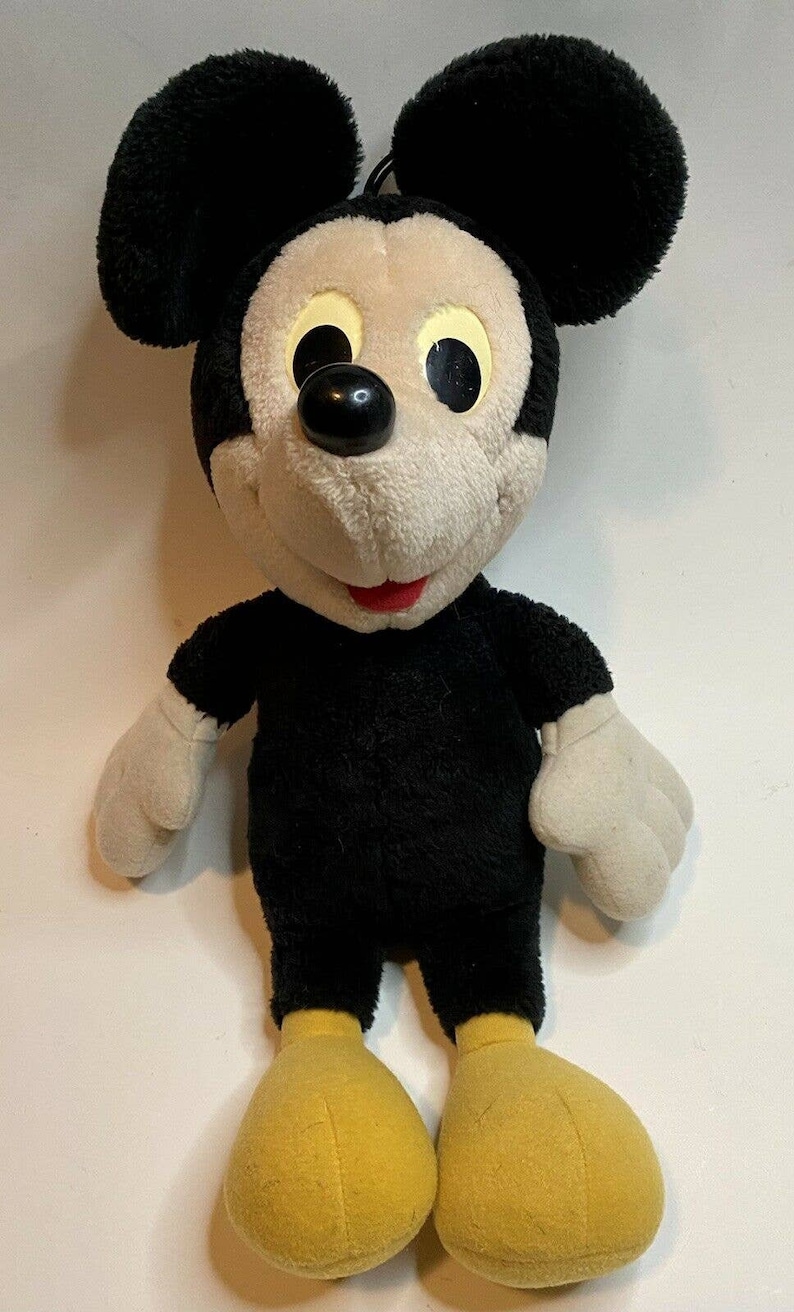 Peut inclure: Une peluche Mickey Mouse avec une fourrure noire, de grandes oreilles noires et un visage cr&egrave;me. Le jouet a un nez noir, des yeux jaunes et une bouche rouge. Il porte une tenue noire avec des gants blancs et des chaussures jaunes.