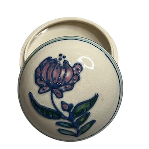 May include: A ceramic trinket box with a lid. The lid features a raised floral design in pink and blue against an off-white background. The box has a light blue and green trim.