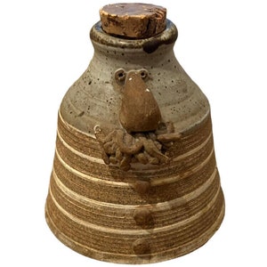 May include: A handmade ceramic bottle with a cork stopper. The bottle is grey with brown horizontal stripes and a whimsical face design. The face has a large nose and an octopus-like design below.