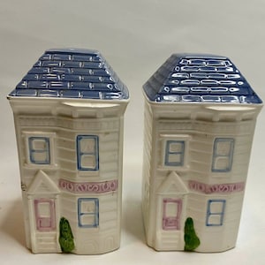 May include: A pair of ceramic salt and pepper shakers shaped like houses. The houses are white with blue roofs, pink and blue window frames, and a green bush. The design is reminiscent of a Victorian style.