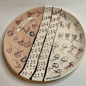 Puede incluir: Un plato decorativo de cerámica con un diseño único. El plato presenta una combinación de colores melocotón claro y blanco roto, con detalles en negro y marrón. El diseño incluye un patrón de rayas diagonales con líneas negras y lunares, y texto manuscrito.