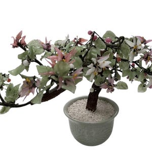 May include: A decorative glass bonsai tree with pink and white flowers and green leaves. The tree is in a gray ceramic pot filled with white pebbles. The branches extend horizontally, creating a balanced aesthetic.