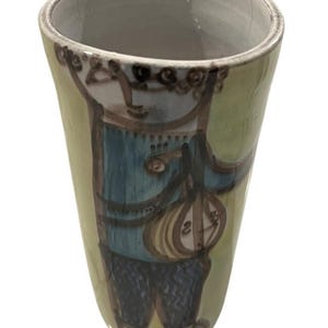 May include: A hand-painted ceramic vase with a whimsical design. The vase features a figure in a blue jacket and dark pants, holding a musical instrument. The background is a light green color. The vase is approximately 5 inches tall.