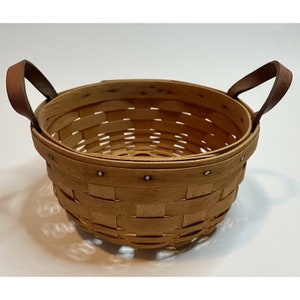 May include: A round, woven basket with a light brown finish. It has two brown leather handles and is secured with small metal rivets. The basket is designed with a classic, rustic aesthetic.