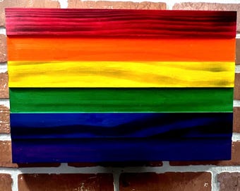 May include: A rectangular wooden wall art piece featuring five horizontal planks painted in a vibrant rainbow gradient. The colors transition from deep red at the top, followed by orange, yellow, green, and a dark blue-purple shade at the bottom.