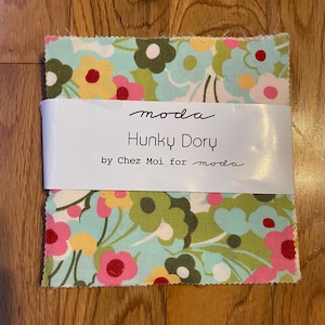 Può includere: Un mucchio di quadrati di tessuto con un motivo floreale rosa, giallo, verde e bianco su sfondo azzurro. Il tessuto è avvolto da un'etichetta bianca che recita "moda Hunky Dory by Chez Moi for moda."