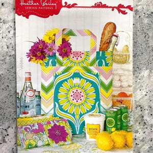 May include: A Heather Bailey sewing pattern package for New Leaf Folding Totes. The cover features a colourful tote bag with a green, yellow, and pink chevron and floral pattern, alongside smaller matching pouches, lemons, and food items.