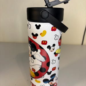 May include: A white cylindrical tumbler featuring a repeating pattern of Mickey Mouse heads, gloves, and shoes. A large circular graphic shows Mickey Mouse, and the name Cynthia is printed in red letters. The tumbler has a black lid with a flip-top spout.
