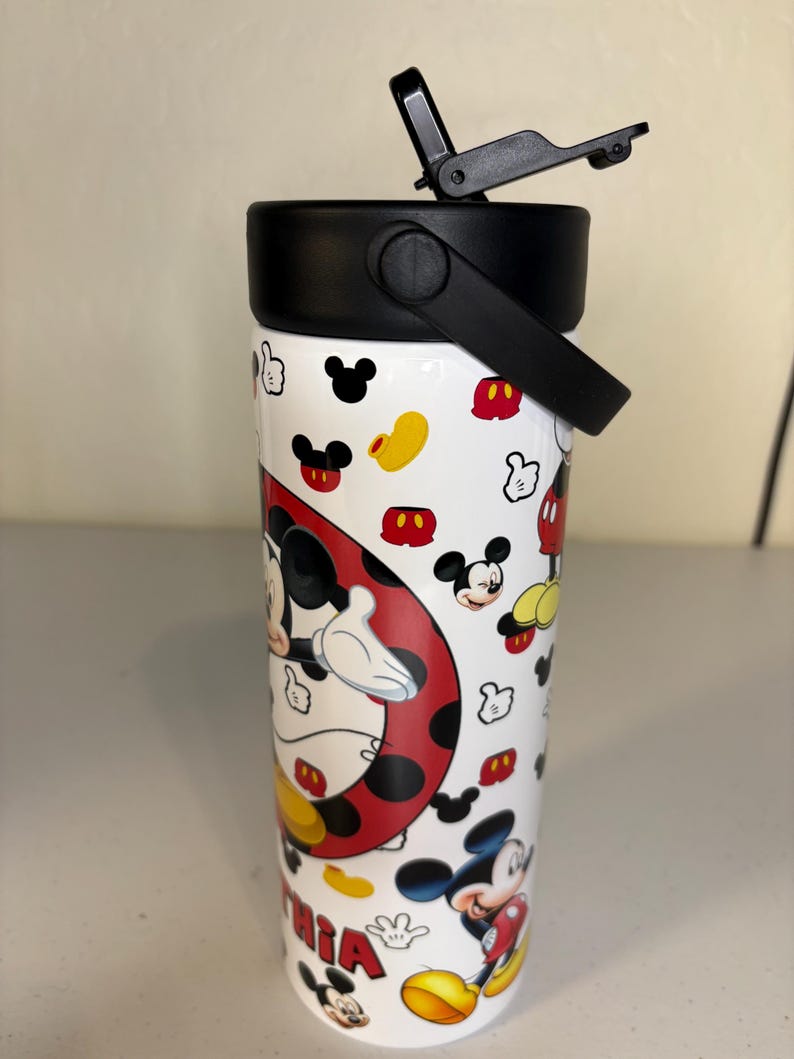 May include: A white cylindrical tumbler featuring a repeating pattern of Mickey Mouse heads, gloves, and shoes. A large circular graphic shows Mickey Mouse, and the name Cynthia is printed in red letters. The tumbler has a black lid with a flip-top spout.