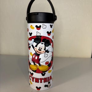 May include: A white cylindrical water bottle with a black lid and handle. The bottle features a repeating pattern of Mickey Mouse icons and a central circular graphic of Mickey Mouse with the name CYNTHIA printed in red capital letters below.