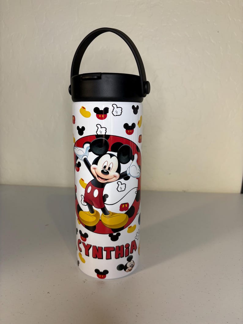 May include: A white cylindrical water bottle with a black lid and handle. The bottle features a repeating pattern of Mickey Mouse icons and a central circular graphic of Mickey Mouse with the name CYNTHIA printed in red capital letters below.
