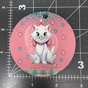 May include: A round, pink ornament featuring a white cartoon cat with a pink bow and accents. The ornament has a glittery top edge and is decorated with flowers and a decorative border. The ornament has a hole at the top for hanging.
