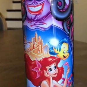 Puede incluir: Un vaso de acero inoxidable con una imagen colorida de Ariel, &Uacute;rsula y Sebasti&aacute;n de la pel&iacute;cula de Disney La Sirenita. El vaso tiene un fondo azul con una imagen morada, verde y amarilla de &Uacute;rsula, una Ariel pelirroja y un Sebasti&aacute;n amarillo.