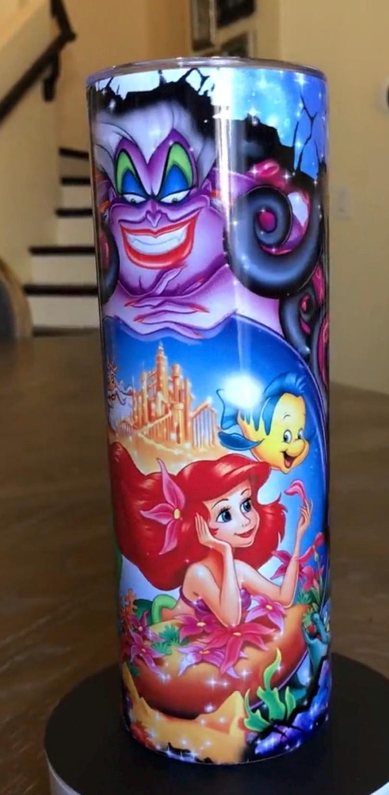 Puede incluir: Un vaso de acero inoxidable con una imagen colorida de Ariel, &Uacute;rsula y Sebasti&aacute;n de la pel&iacute;cula de Disney La Sirenita. El vaso tiene un fondo azul con una imagen morada, verde y amarilla de &Uacute;rsula, una Ariel pelirroja y un Sebasti&aacute;n amarillo.