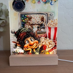 May include: A decorative light with a whimsical design. The image features a smiling elf, a vintage television showing two girls, and a popcorn bucket. The top of the light has the text "North Pole Surveillance Camera". The light sits on a wooden base.