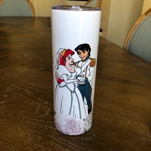 Puede incluir: Un vaso de acero inoxidable blanco con un gr&aacute;fico de Ariel y el Pr&iacute;ncipe Eric de La Sirenita en su atuendo de boda. El gr&aacute;fico incluye detalles florales alrededor de la parte inferior del vaso.
