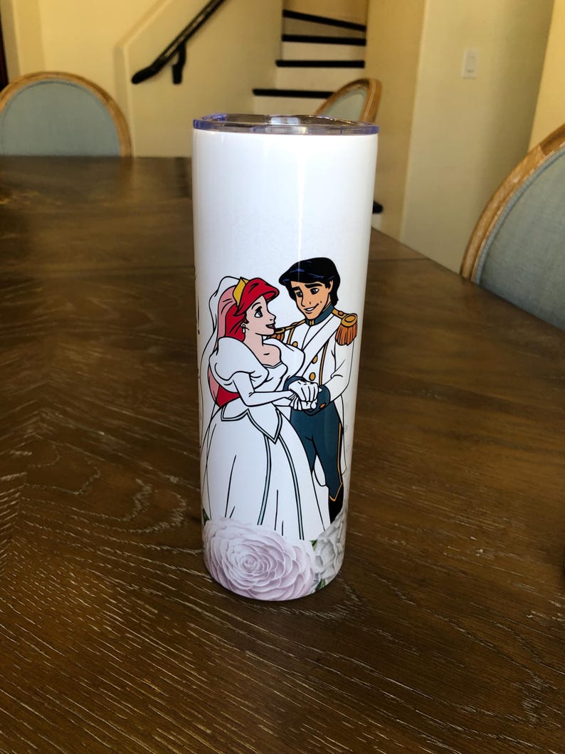 Puede incluir: Un vaso de acero inoxidable blanco con un gr&aacute;fico de Ariel y el Pr&iacute;ncipe Eric de La Sirenita en su atuendo de boda. El gr&aacute;fico incluye detalles florales alrededor de la parte inferior del vaso.