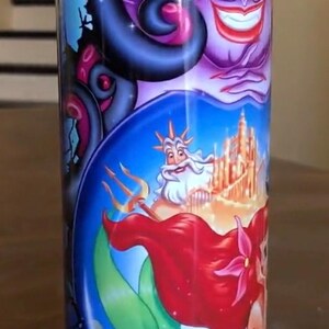 Puede incluir: Un vaso de acero inoxidable colorido con un gr&aacute;fico de &Uacute;rsula, Ariel y el rey Trit&oacute;n de la pel&iacute;cula de Disney La Sirenita. El vaso presenta una gama de colores azul, verde, morado y rojo.