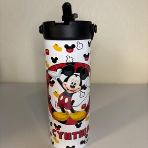 May include: A white cylindrical water bottle with a black lid and a flip-up straw. The bottle features a repeating pattern of Mickey Mouse icons and a central graphic of Mickey Mouse with the name CYNTHIA printed in red block letters below.