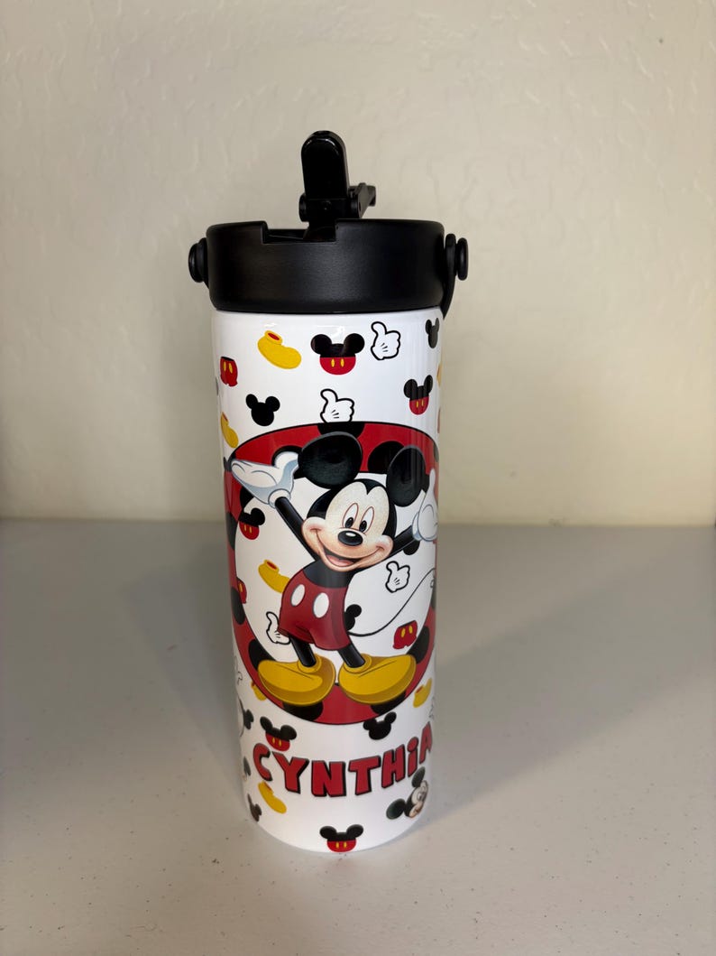 May include: A white cylindrical water bottle with a black lid and a flip-up straw. The bottle features a repeating pattern of Mickey Mouse icons and a central graphic of Mickey Mouse with the name CYNTHIA printed in red block letters below.