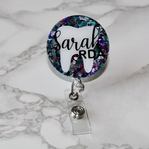 Puede incluir: Un portagafete blanco en forma de diente con un fondo transparente y un diseño de purpurina azul, morado y rosa. El nombre "Sarah RDA" está escrito en letras negras en el portagafete.