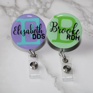 May include: Two personalized badge reels with clear clips. The left reel is purple with a teal letter "E" and the text "Elizabeth DDS". The right reel is green with a green letter "B" and the text "Brooke RDH".