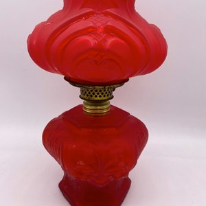 May include: A red glass oil lamp with a decorative design. The lamp has a round, fluted top and a hexagonal base.