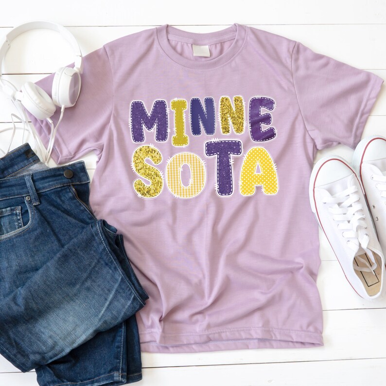 K&ouml;nnte beinhalten: Ein lilafarbenes T-Shirt mit dem Wort "MINNESOTA" in einem Patchwork-Design. Die Buchstaben sind in goldfarbenem Glitzer, Lila und gelben Mustern gehalten. Das Shirt wird mit blauen Jeans, wei&szlig;en Turnschuhen und Kopfh&ouml;rern kombiniert.