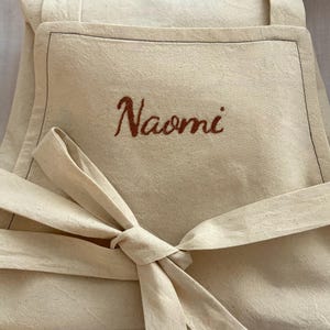 Puede incluir: Delantal color crema con el nombre "Naomi" bordado en una fuente marrón. El delantal tiene un bolsillo delantero y una cintura atada. La tela parece ser de algodón o lino natural sin blanquear.